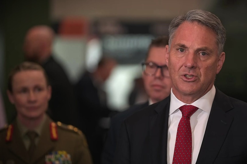 Richard Marles at a press conference wearing a black suit jacket and dark red tie, mid-speech.