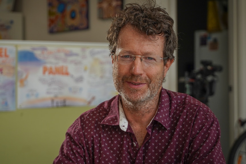 Jonathan Pilbrow looks at the camera wearing spectacles and a maroon shirt.