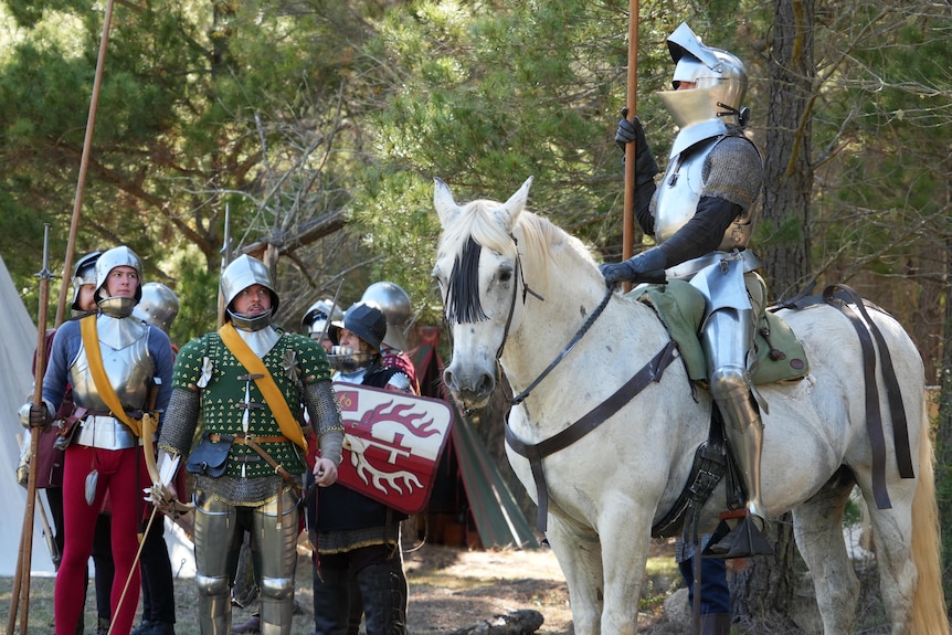 Men in armor stand next to a knight on horseback .