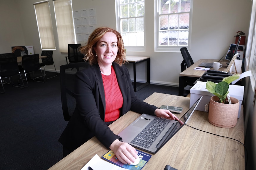 A woman in a red top and black jacket sits at a laptop in an office. 