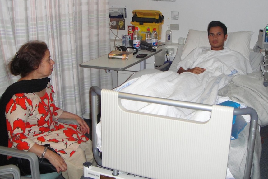 Usman Khawaja seated in a hospital bed in a ward. His mother sits in a chair at the foot of the bed.