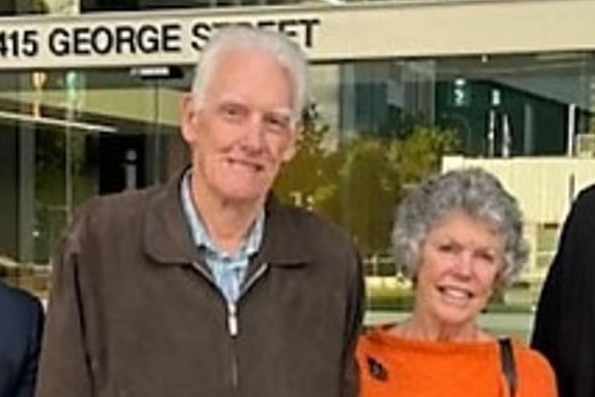 An white-haired man in a brown cardigan stands smiling next to a shorter grey-haired woman in an orange top outside court.