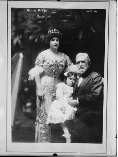 David Mitchell holds Nellie Melba's young niece while Nellie stands next to them wearing an ornate gown.