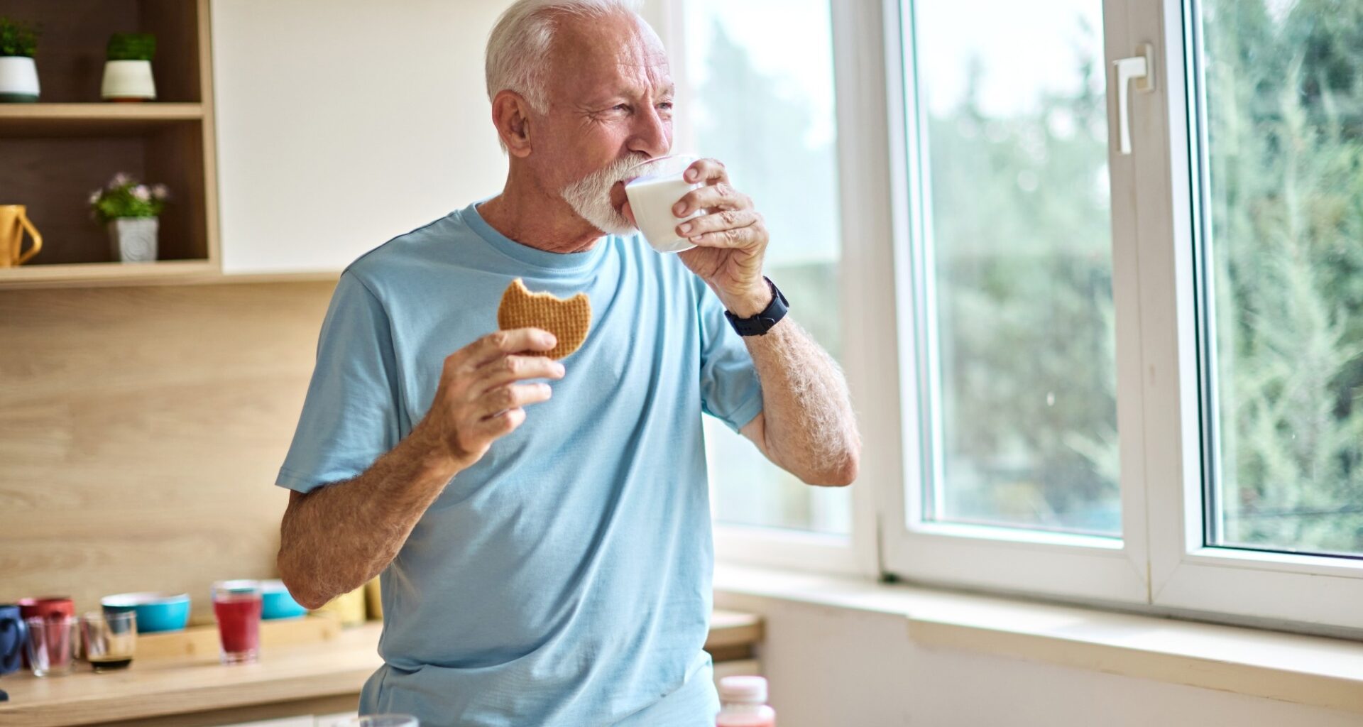 A senior man is eating a cokie energy bar and drinking milk in kitchen.