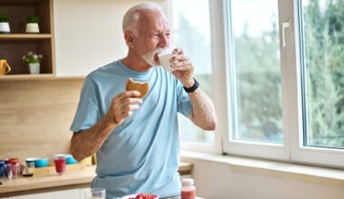 A senior man is eating a cokie energy bar and drinking milk in kitchen.