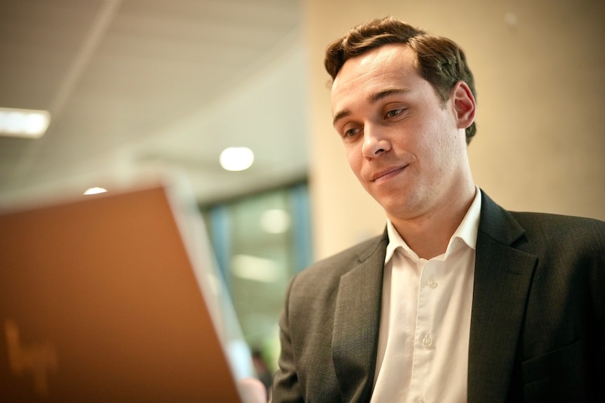 Lochlan Halloway, wearing a suit, looks at a screen in an office.