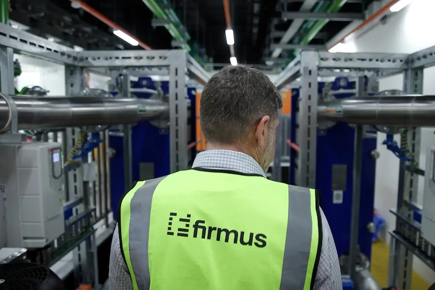 A man in a fluro vest with the word Firmus on it walks past shelves of computer equipment.
