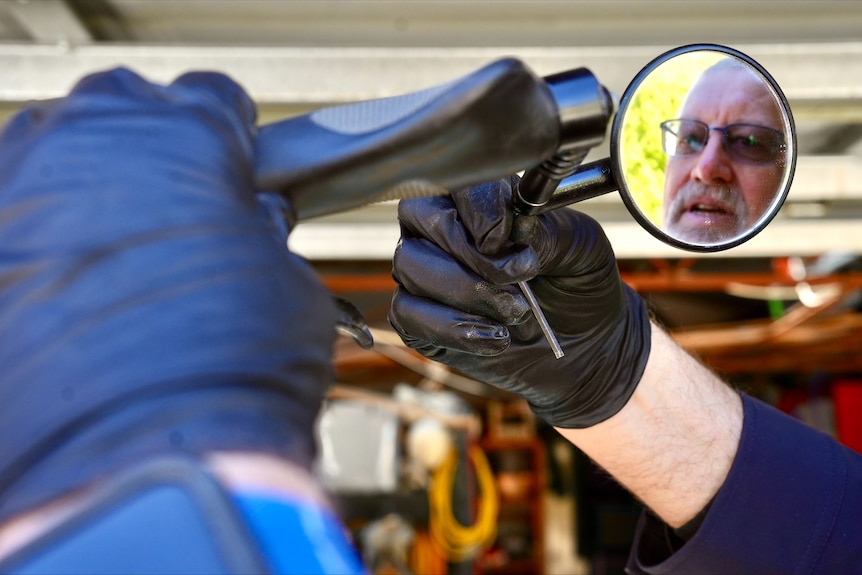 A man with short grey hair and glasses in the side mirror of a motorcycle as he fixes it.