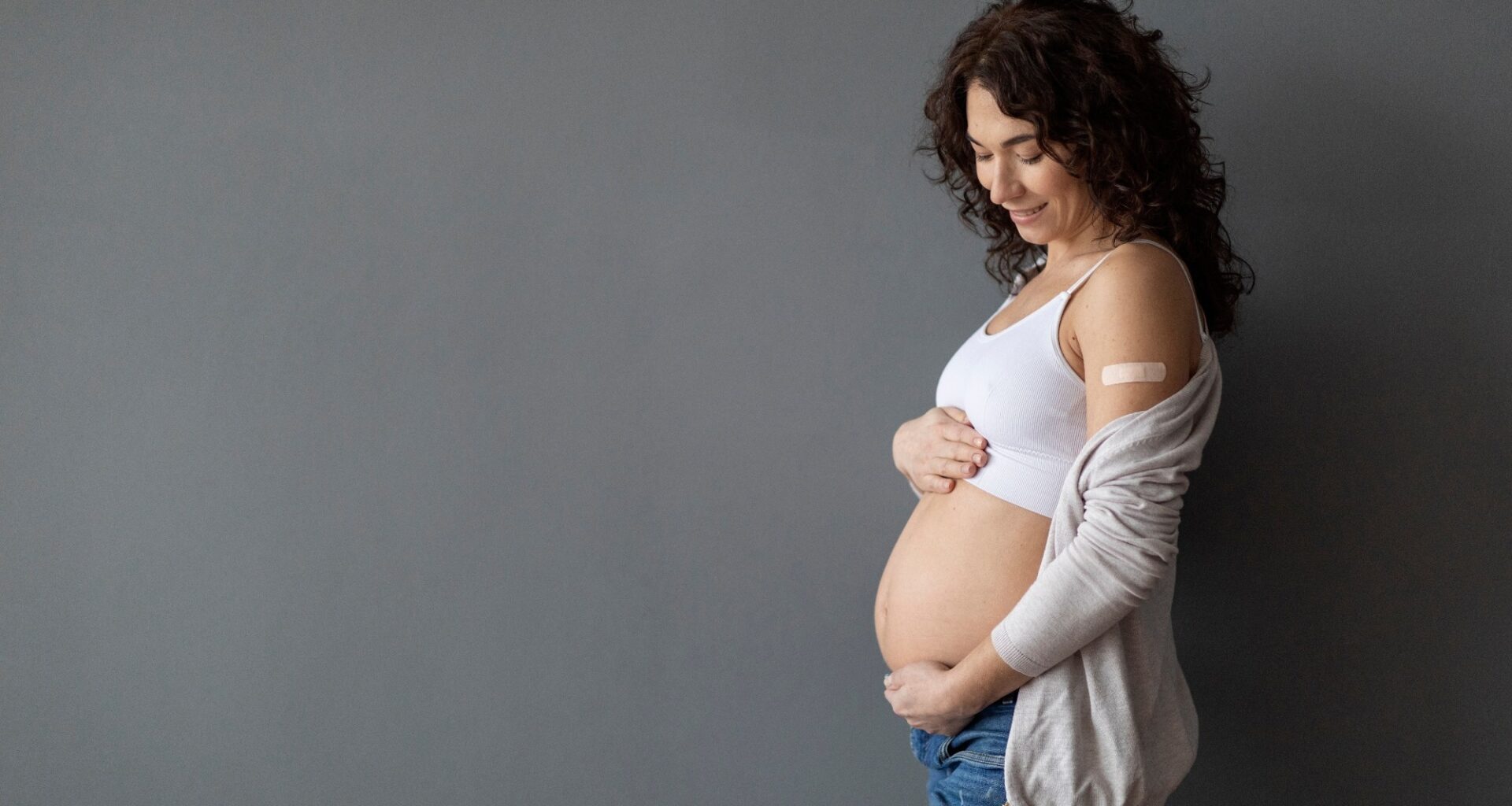 Happy Pregnant Female With Adhesive Bandage On Shoulder Embracing Her Belly While Standing Over Grey Studio background,