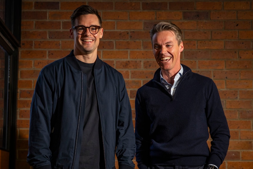 two men in dark tops stand against a red brick wall, one brunette and taller, the other with grey hair
