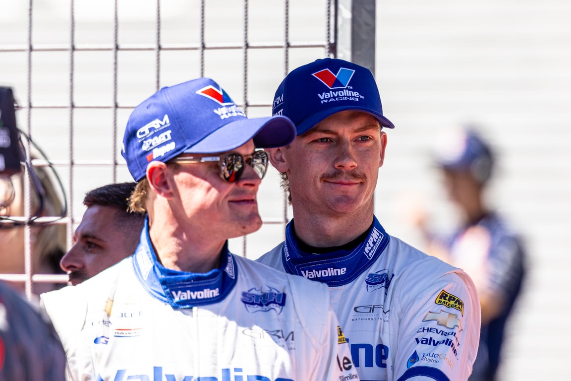 Two drivers in Valvoline Racing suits and caps stand together in the pit area.