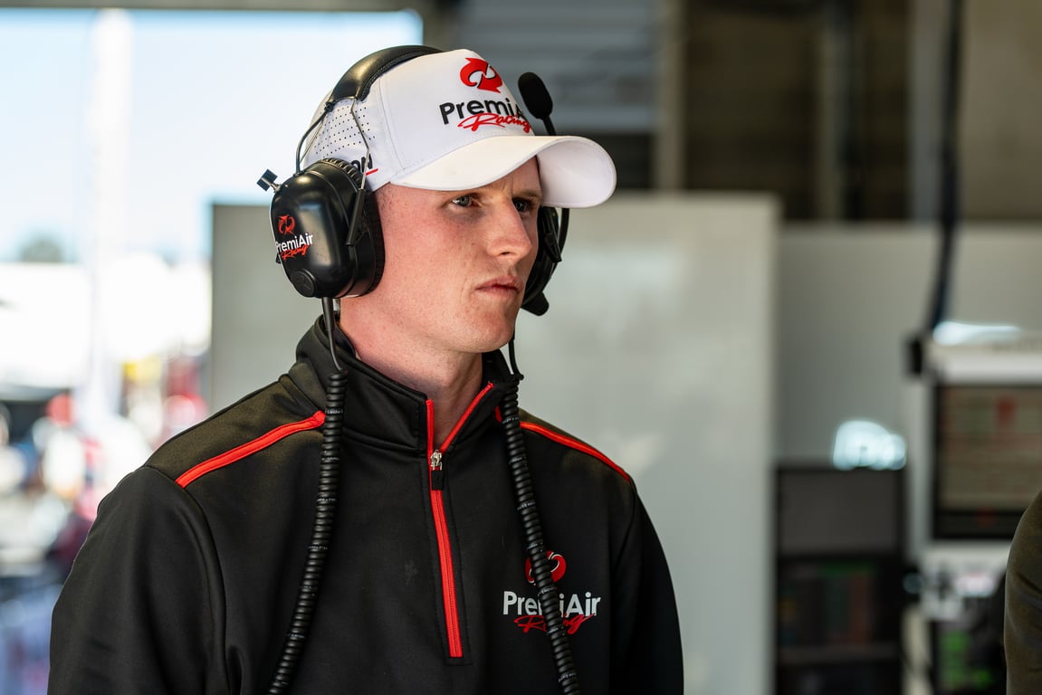 Male race crew member in white PremiAir Racing cap and headset standing in a blurred pit garage.