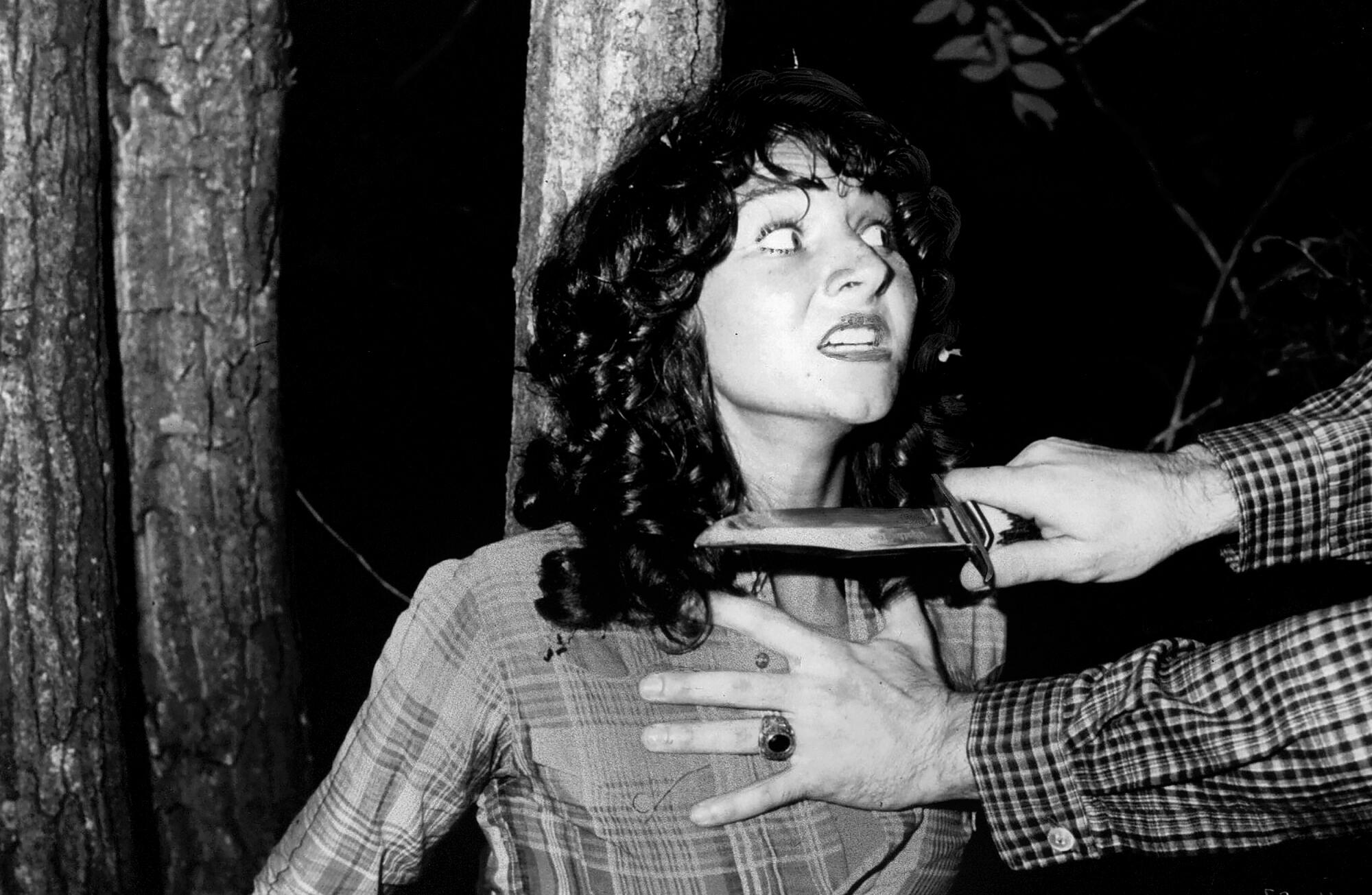 A woman standing against a tree with a knife being held to her throat by a person with only their hands visible.
