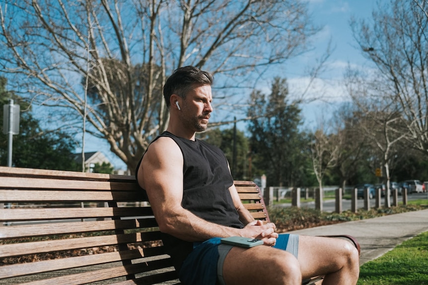 Muscular man in dark single, brightly-coloured shorts, on park bench with in-ear headphones