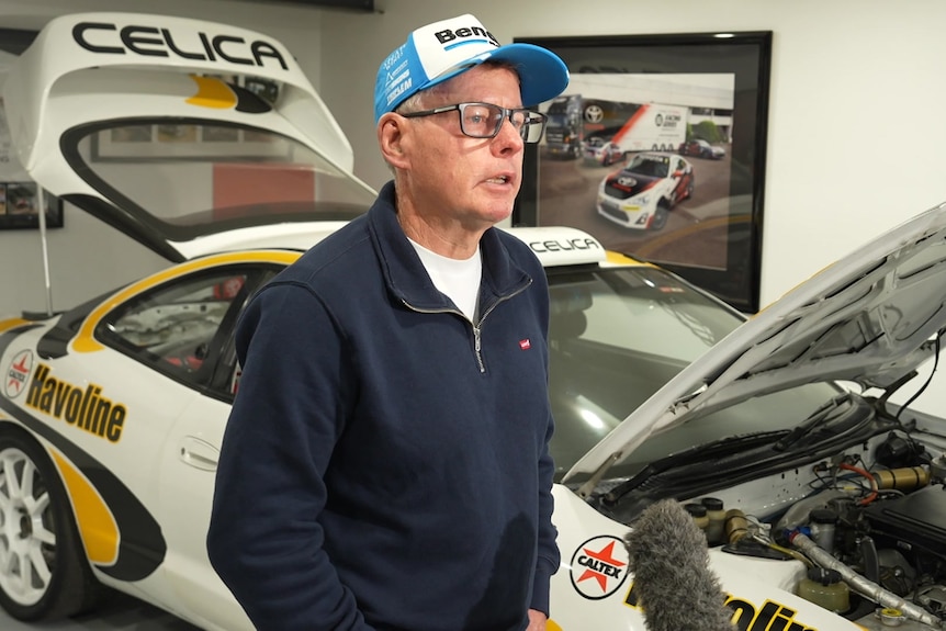 A man wearing a motorsports cap stands in front of a supercar speaking.