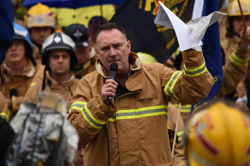 A group of firefighters in tan coats, one holding a microphone.