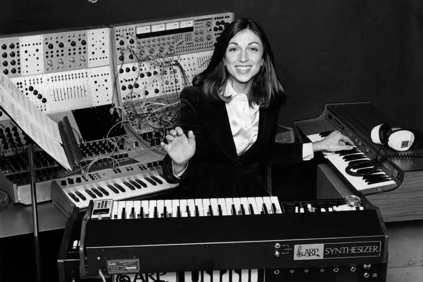 Black and white photo of young woman in business suit sat among bank of keyboards and modular synthesizers