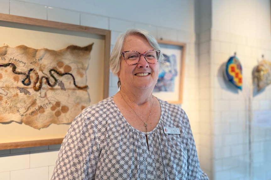 A woman with grey hair and glasses stands in an art gallery displaying embroidery. 