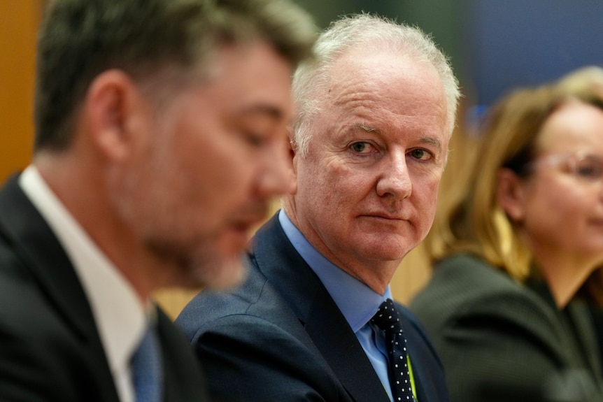 A grey-haired man (Hugh Marks) wearing a suit is in focus, with blurred smartly dressed people sitting either side.