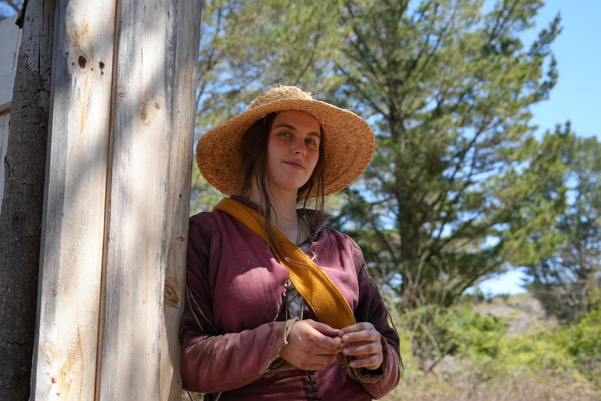 A woman wearing a red tunic and a large straw hat smiles shyly down at the camera 