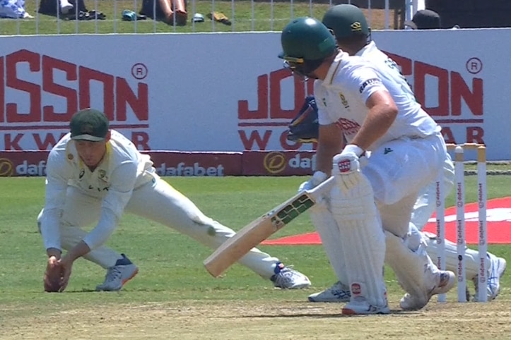 A cricketer takes a low catch with the ball hitting grass through his fingers.