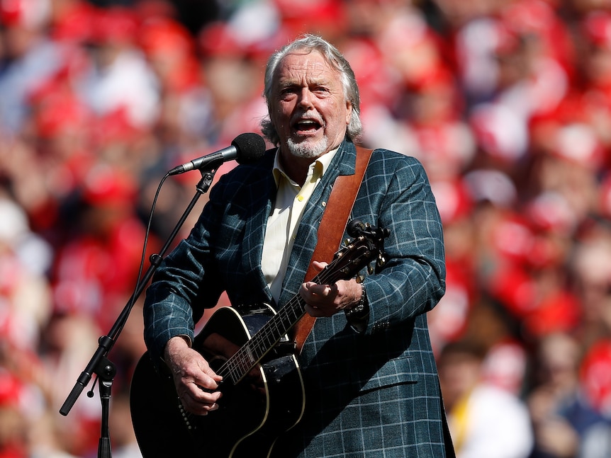 Mike Brady strums a guitar and sings into a microphone.
