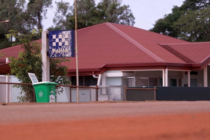 A low-set building with a police sign next to it. In the foreground is a road covered by red dirt.