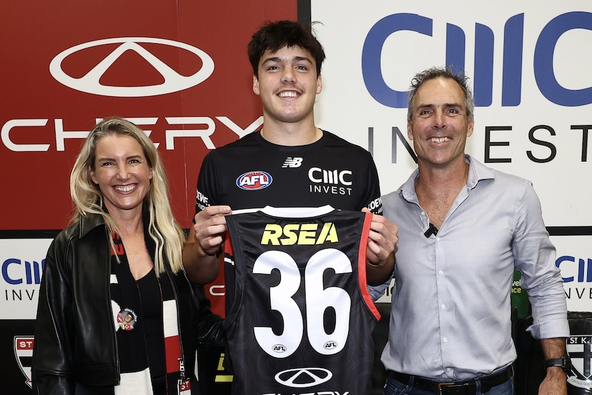 Charlie Banfield smiles and holds the St Kilda jumper #36, flanked by his mother and father in the St Kilda rooms.