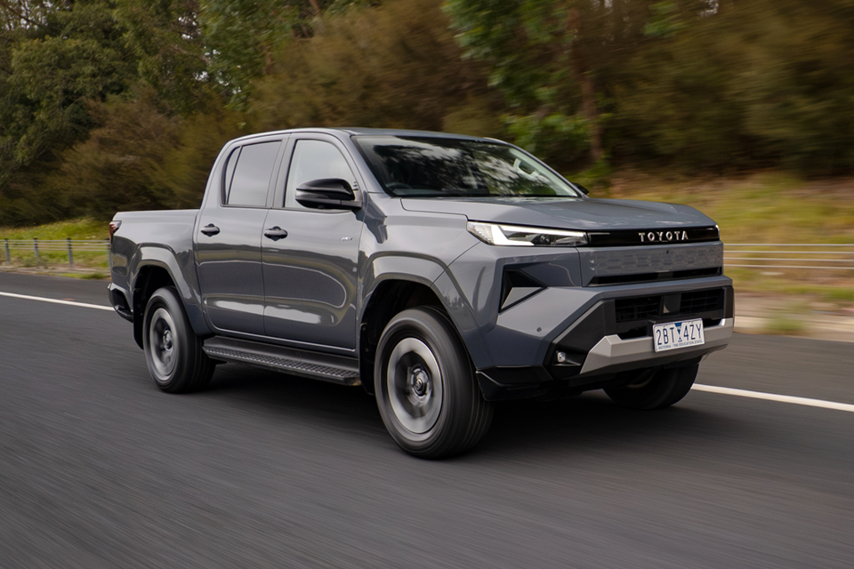 A grey 2026 Toyota HiLux BEV double-cab ute driving on a road, seen from the front-left side