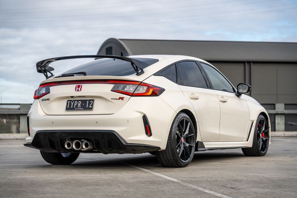 A white Honda Civic Type R hatchback seen from the rear three-quarter angle in a car park, showing its large rear wing, triple exhaust outlets, and red-accented taillights.