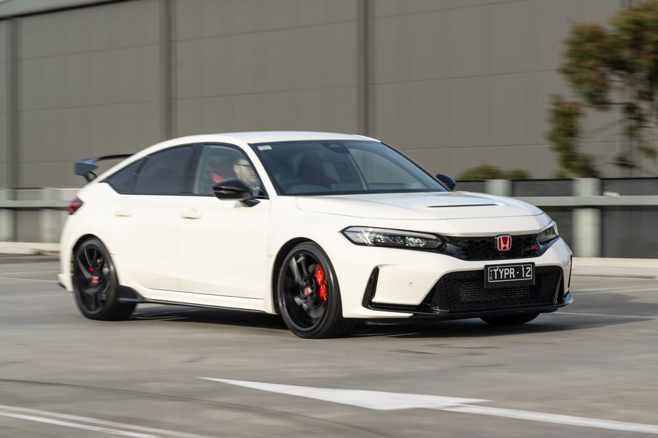 A white Honda Civic Type R hatchback driving through a car park, seen from the front three-quarter angle