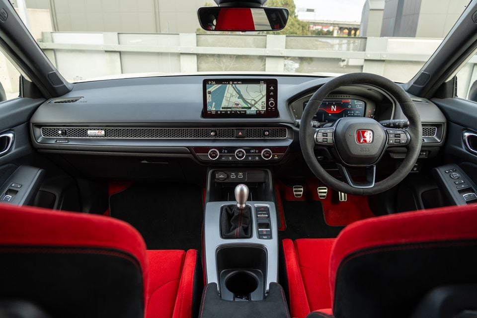 Interior of a Honda Civic Type R showing the red and black cabin with flat-bottom steering wheel, touchscreen navigation, manual gear shifter, and digital instrument cluster