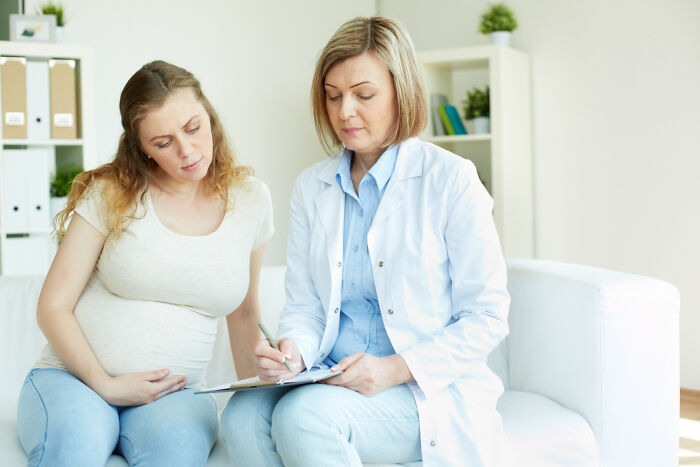 A female doctor writing on a clipboard while consulting with a pregnant patient.