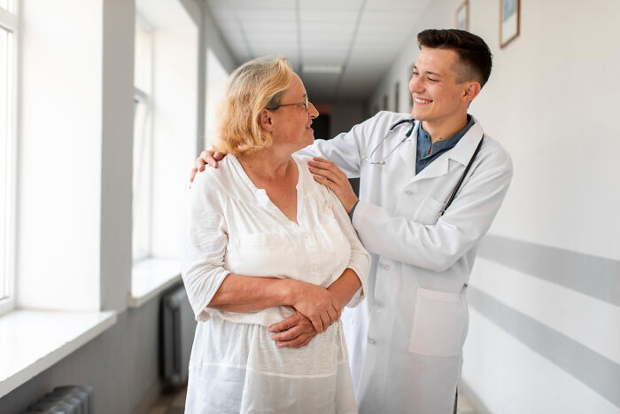 Young smiling male doctor walking down a hospital hallway with an elderly female patient.