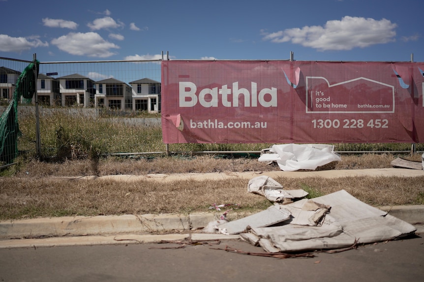A red hoarding with "Bathla" on it sits on rough ground with broken plasterboard strewn across the road.