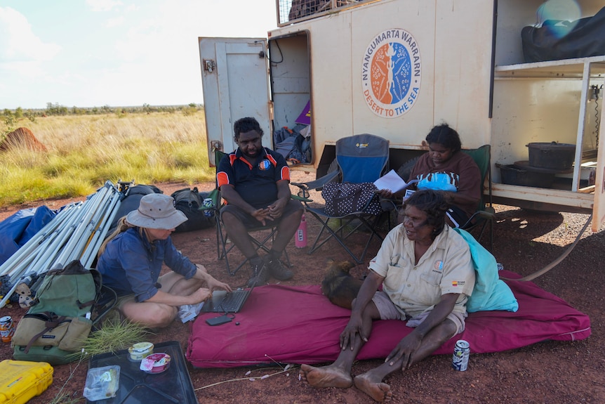 A group of Aboriginal rangers and a Central Queensland university researcher sit in a circle behind a trailer in the desert.