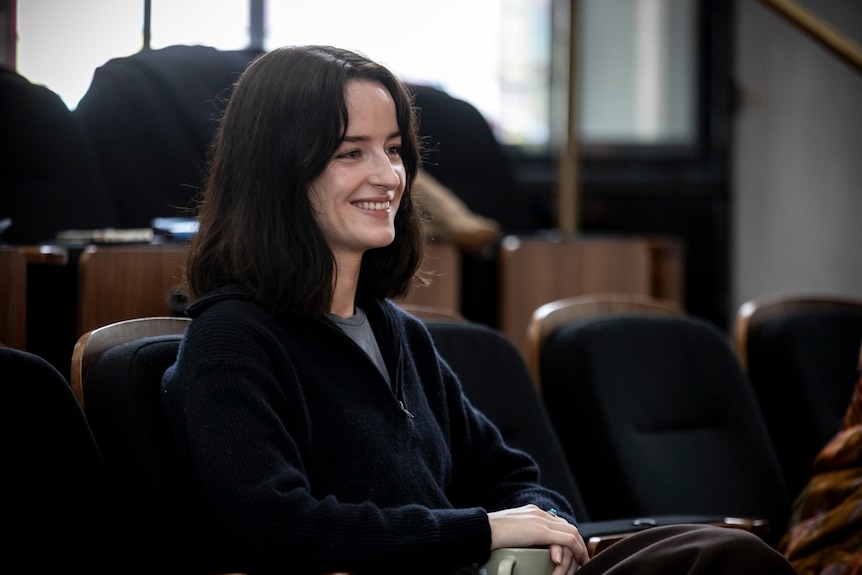 In a rehearsal room, Grace Chapple, early 30s, wearing a cardigan, smiles, sitting in a seating bank.