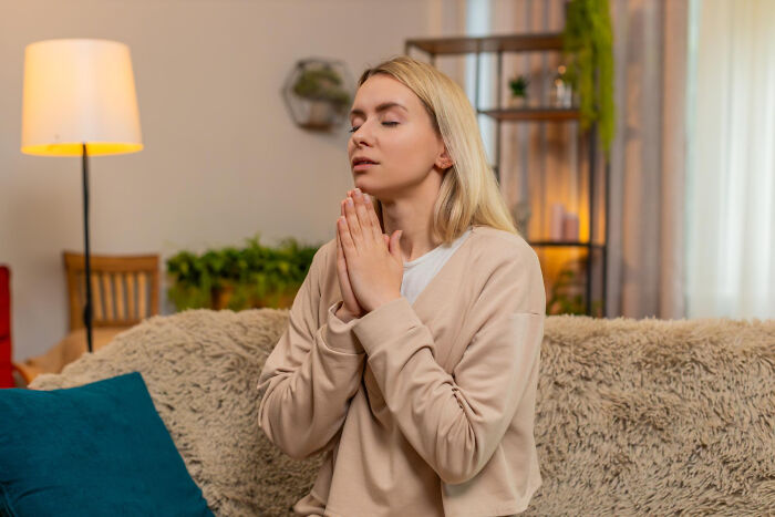 A young woman sitting on a couch with eyes closed and hands clasped in prayer or deep thought
