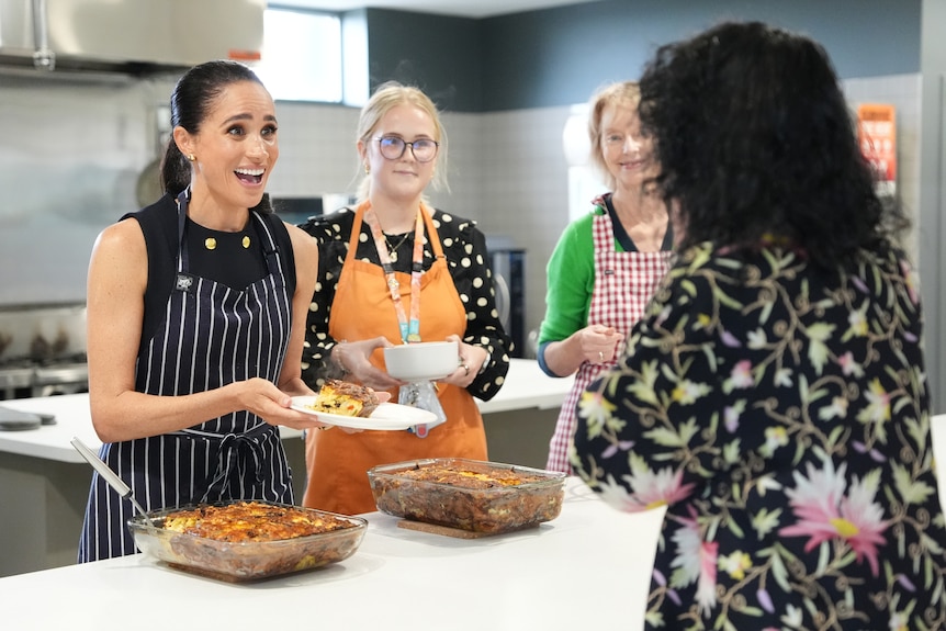 A dark-haired woman (Meghan Markle) serving lunch alongside other women in aprons.