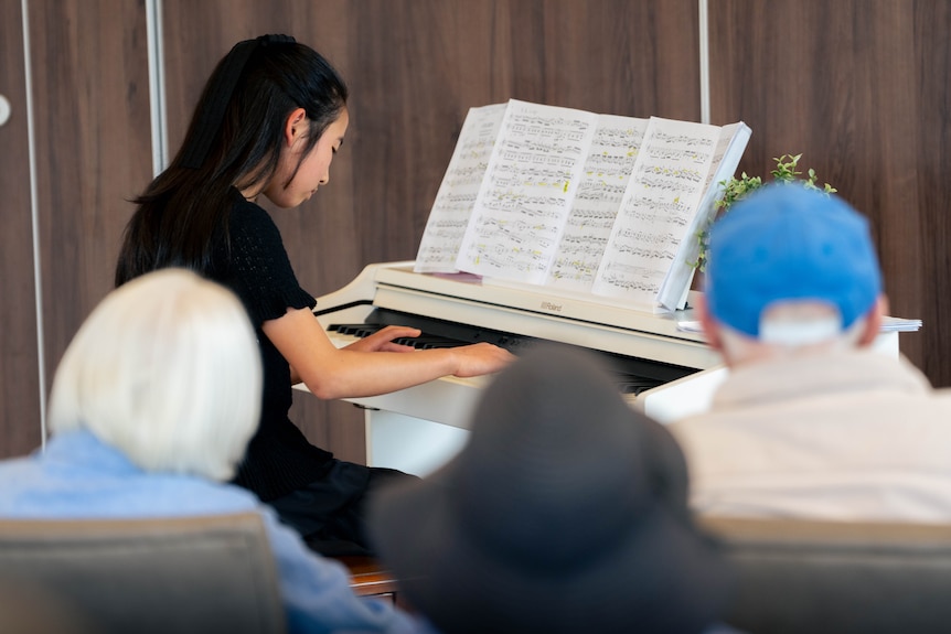 Min, in a black dress, performs a piano piece on a white piano of aged care residents.