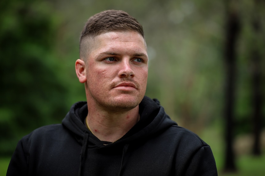 Man wearing black hoodie with short cut hair stands looking sideways in green bushland