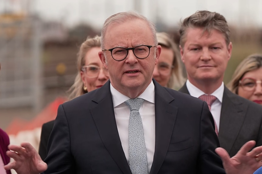 A white haired man with dark rimmed glasses in a dark suit, white shirt and pale green tie stands in front of a group of people.