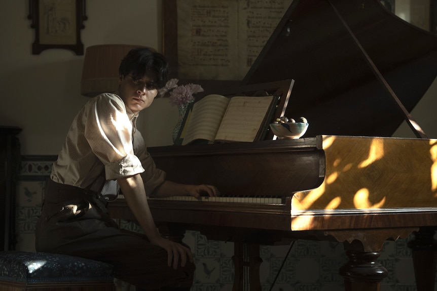 A young man with black hair wearing a white shirt slumps at a piano