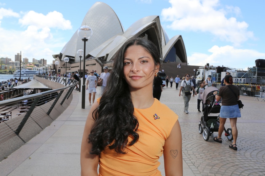 Daniella Gilboa outside the Sydney Opera House, with dark hair, an orange shirt, and a bee brooch.