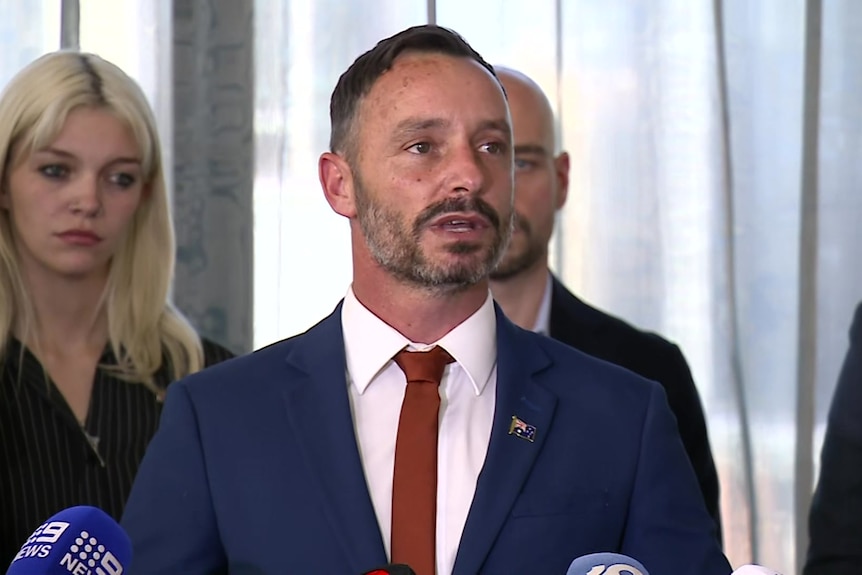A man in a blue suit with a red tie stands in front of microphones for a press conference.