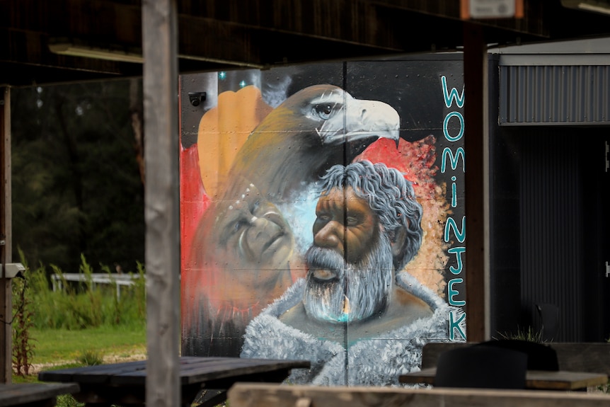 A colourful wall mural with Indigenous icongraphy and the word Wominjeka is displayed on the wall of a building in a yard