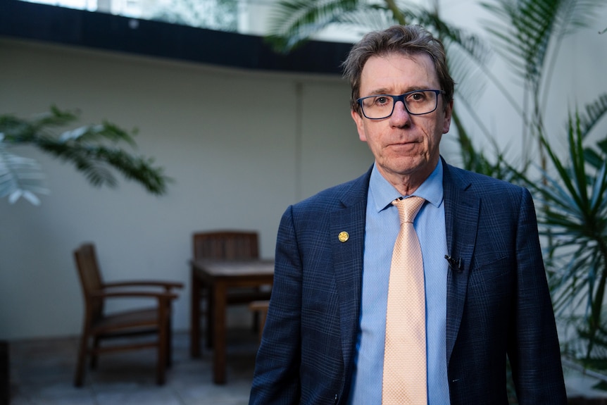 A middle aged man wearing a suit and tie, wearing glasses, looking serious.