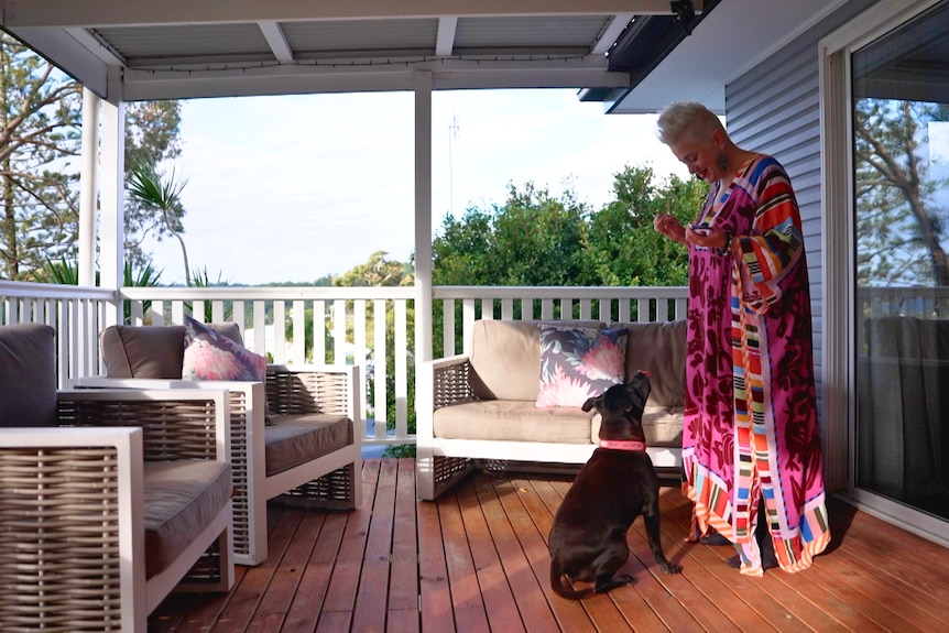 Katie Noonan with short blonde hair wears a colourful kaftan standing on a outdoor home deck looking down at her staffy dog.