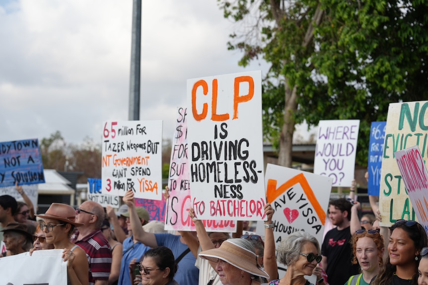 A person holds a sign reading "CLP is driving homelessness (to Batchelor)" while surrounded by other protestors.
