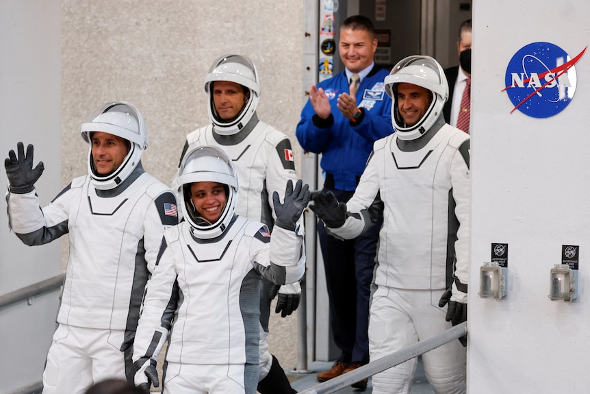 Three male and one female astronaut seen walking and smiling in white spacesuits down a ramp.